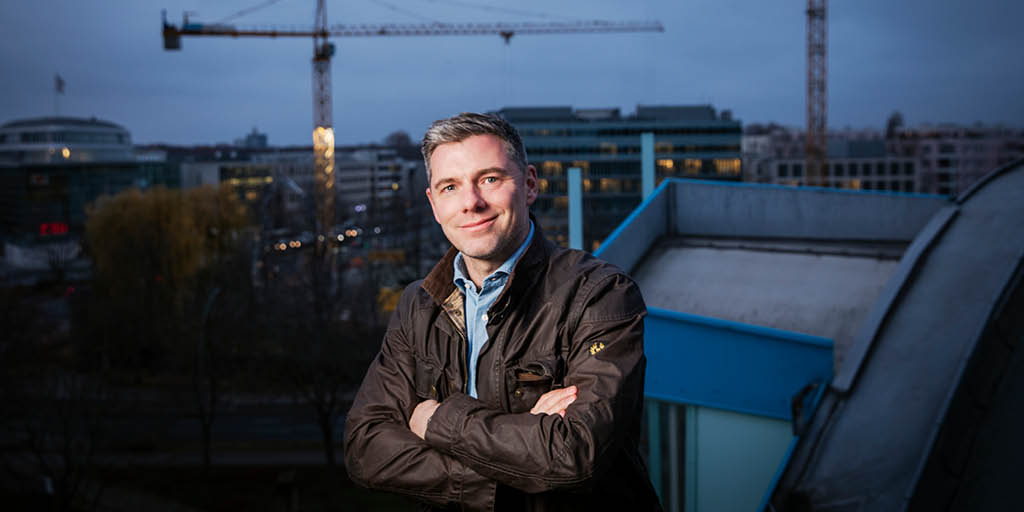 Tobias Beuler steht lächelnd mit verschränkten Armen auf einer Dachterrasse, im Hintergrund sind Baukräne und moderne Bürogebäude bei Abenddämmerung zu sehen.